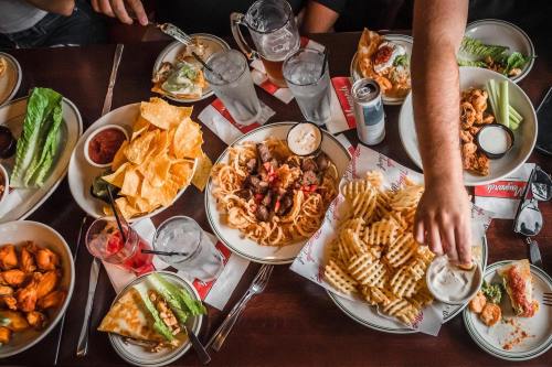 A table filled with various dishes, including nachos, pasta, chicken wings, and drinks, ready for sharing.