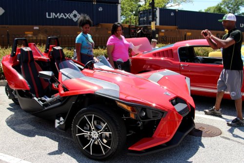 A red three-wheeled vehicle is parked, with people admiring it and taking photos in a sunny outdoor setting.