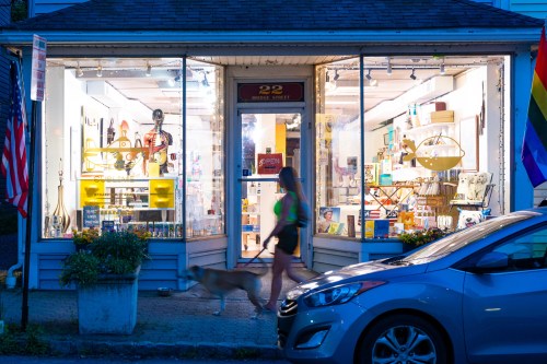 A woman walks a dog past a brightly lit shop window displaying various items, with flags and a parked car nearby.