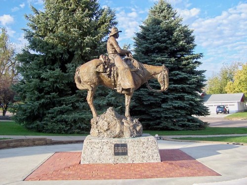 A bronze statue of a cowboy on horseback, mounted on a stone pedestal, surrounded by trees and a clear sky.