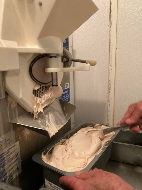 A person scoops creamy ice cream from a machine into a container, with the machine visible in the background.