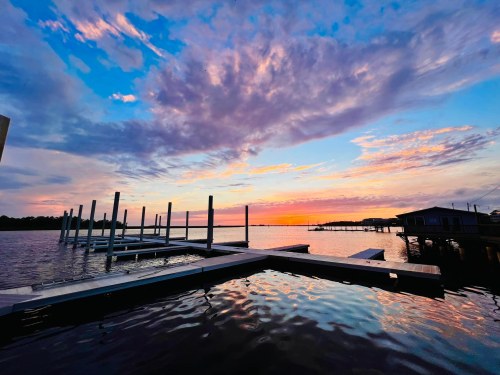Sunset over a calm waterway, with docks and clouds reflecting vibrant colors in the sky.