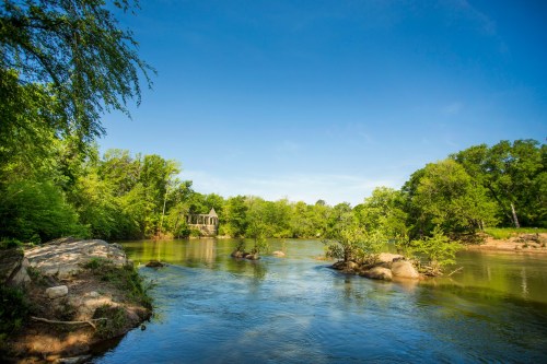 A serene river scene with lush greenery, rocks, and a clear blue sky, featuring a house in the background.
