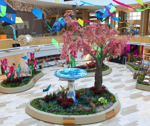 Colorful indoor garden featuring a large cherry blossom tree, vibrant flowers, butterflies, and decorative kites overhead.