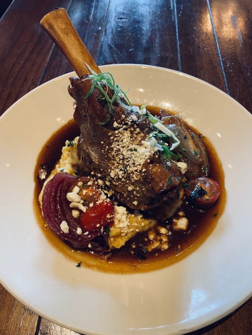 A plated dish featuring a lamb shank on creamy polenta, garnished with green onions and cherry tomatoes in a rich sauce.