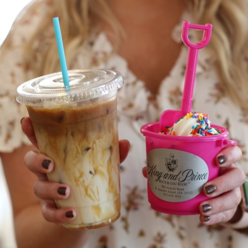 A person holds a cold coffee drink and a pink bucket filled with ice cream and sprinkles, featuring a small shovel.