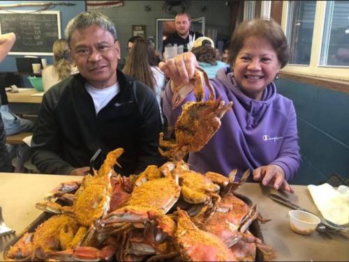 A man and woman smile while holding a crab above a large pile of seasoned crabs on a table in a casual dining setting.