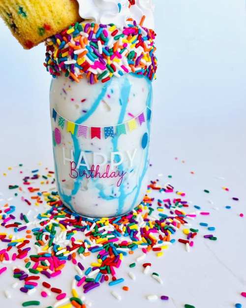 A festive birthday milkshake in a jar, topped with whipped cream, sprinkles, and a piece of cake. Colorful decorations surround it.