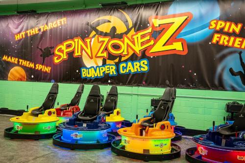 Colorful bumper cars lined up in front of a vibrant "Spin Zone Z" banner in an amusement area.