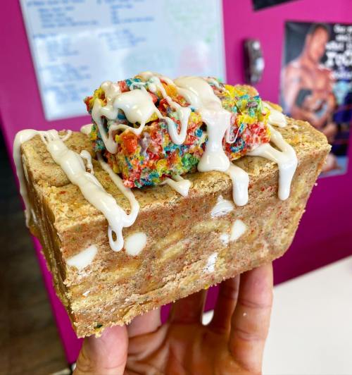 A hand holding a colorful dessert bar topped with white drizzle, set against a vibrant pink background.