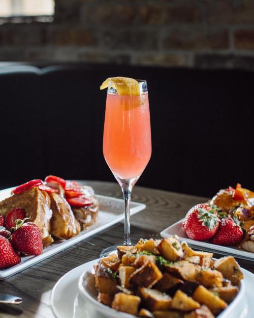 A vibrant cocktail with a lemon slice stands tall among plates of French toast and seasoned potatoes, garnished with strawberries.