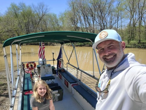 A man and a girl smile on a boat by a muddy river, surrounded by trees on a sunny day.
