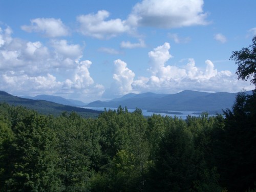 A scenic view of mountains and a lake, surrounded by lush green trees under a blue sky with fluffy white clouds.