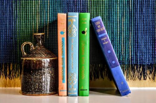 A collection of colorful books beside a dark ceramic jug, set against a textured blue and green woven backdrop.