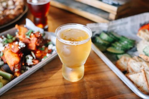 A glass of beer sits on a wooden table, surrounded by plates of wings, cucumbers, and snacks.