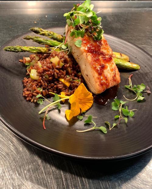 A beautifully plated dish featuring grilled fish, asparagus, wild rice, and garnished with microgreens and an edible flower.