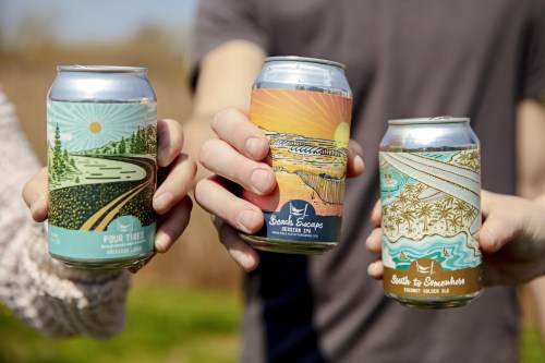 Three hands hold colorful beer cans with scenic designs, set against a sunny outdoor background.