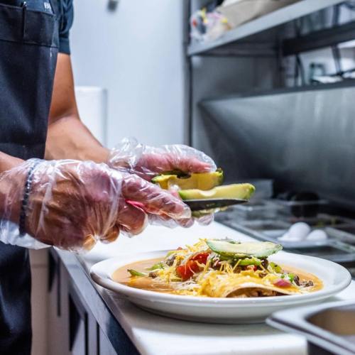 A chef in gloves adds avocado slices to a colorful plate of tacos in a kitchen setting.