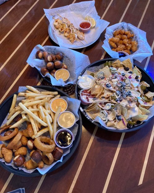 A table spread with various appetizers: nachos, fries, onion rings, chicken skewers, and tater tots with dipping sauces.