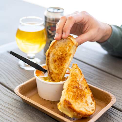 A hand holding a grilled cheese sandwich above a bowl of soup, with a glass of beer nearby on a wooden table.