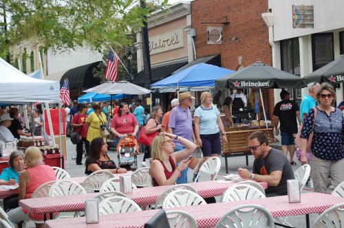 A bustling street fair with people browsing tents, enjoying food, and socializing under colorful canopies.