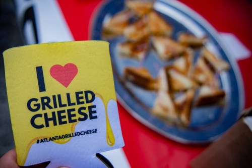 A hand holds a "I Love Grilled Cheese" koozie in front of a plate of grilled cheese sandwich triangles.