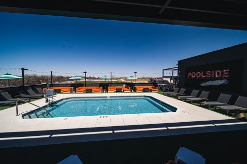 Rooftop pool area with lounge chairs, umbrellas, and a clear blue sky in the background. "POOLSIDE" sign visible.