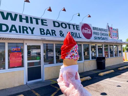 A hand holds a red-iced ice cream cone with sprinkles in front of the White Dotte Dairy Bar & Grill.