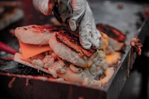 A hand in a glove lifts a grilled sandwich filled with macaroni and cheese and melted cheese on a griddle.