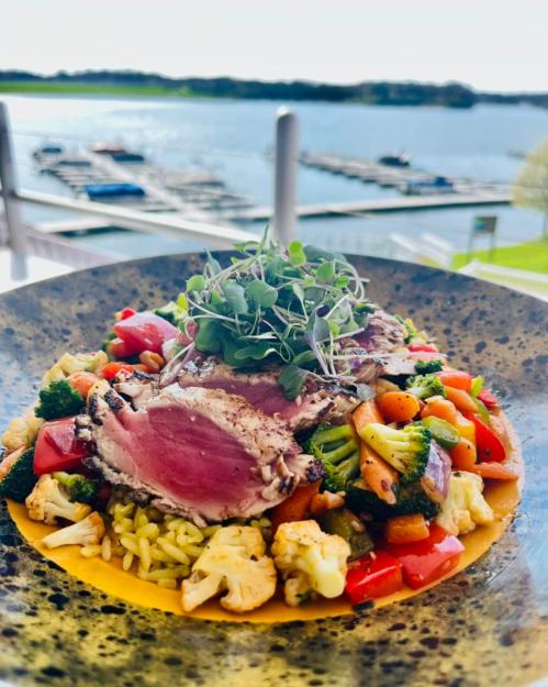 A colorful plate of rice and vegetables topped with sliced grilled meat, with a scenic lake view in the background.