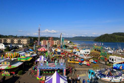 A vibrant carnival scene by the water, featuring colorful rides, tents, and a clear blue sky.