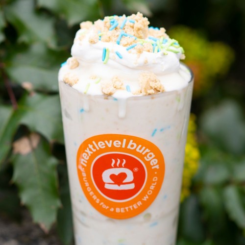 A tall milkshake topped with whipped cream, colorful sprinkles, and cookie crumbles, featuring a logo at the base.