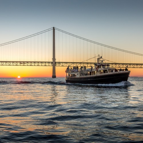 A boat sails on calm waters at sunset, with a bridge silhouetted against the colorful sky.