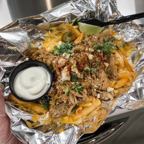 A foil tray filled with loaded fries topped with pulled pork, cheese, jalapeños, cilantro, and a side of sour cream.