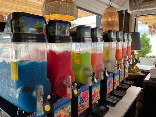 A row of colorful slushie machines with various flavors, including blue, green, red, and white, in a bright setting.