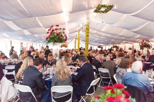 A large indoor event under a tent, filled with guests dining at tables adorned with flowers and elegant decor.