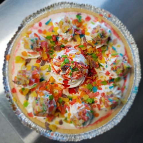A colorful dessert pie topped with whipped cream, fruit cereal, and drizzled with syrup.