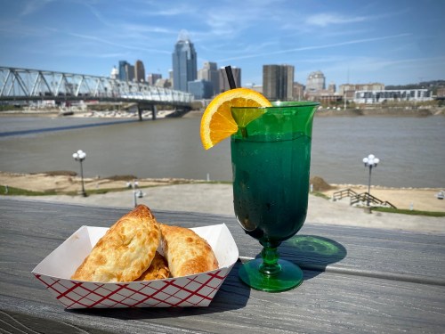 A green drink with an orange slice beside a basket of pastries, with a city skyline and river in the background.