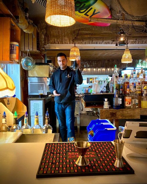 A bartender skillfully mixes drinks at a lively bar, surrounded by bottles and a vibrant tropical atmosphere.