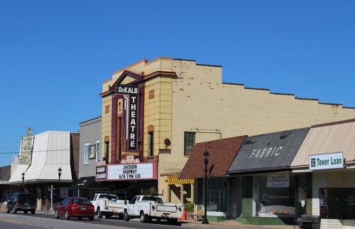 Historic DeKalb Theatre with marquee, surrounded by small shops and clear blue sky. Cars parked along the street.