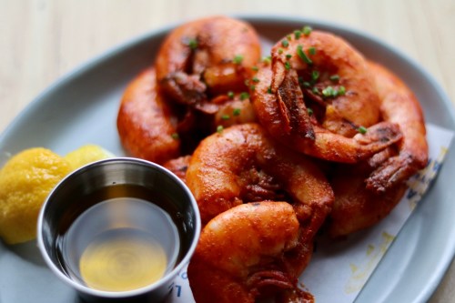 A plate of golden-brown shrimp garnished with herbs, accompanied by a small bowl of dipping sauce and lemon wedges.