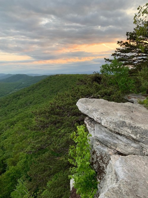 A rocky cliff overlooks lush green mountains under a cloudy sky at sunset, with hints of orange and pink in the horizon.