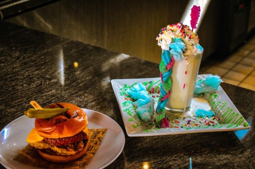 A colorful milkshake topped with whipped cream and sprinkles next to a burger with cheese and pickles on a plate.