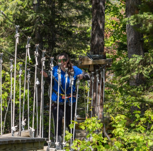 A person in a blue shirt crosses a rope bridge surrounded by trees, wearing sunglasses and safety gear.