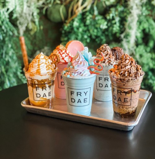 A tray of five colorful milkshakes topped with whipped cream and various decorations, set against a green backdrop.
