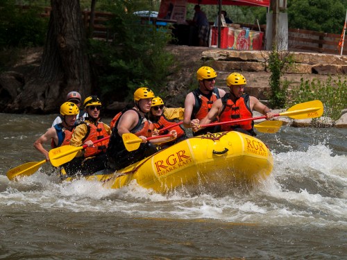 A group of six people in yellow helmets and life jackets paddling a bright yellow raft through whitewater rapids.