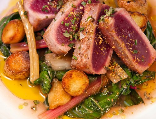 Seared tuna slices on a bed of sautéed greens and roasted potatoes, drizzled with olive oil and garnished with herbs.