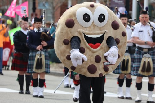 A person in a large cookie costume points while a bagpipe band performs in the background during a parade.
