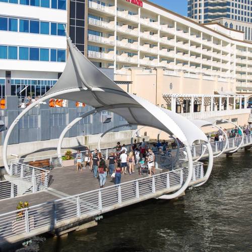 A waterfront promenade with a large canopy, people walking, and buildings in the background on a sunny day.