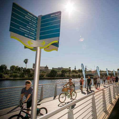 A sunny riverside path with a directional sign, cyclists, and pedestrians enjoying the day.